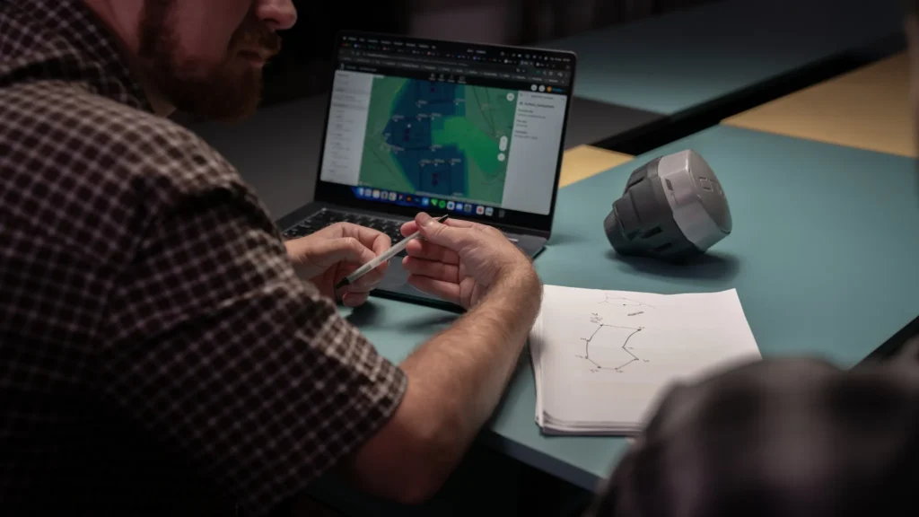 Engineer reviewing GNSS survey data on a laptop while planning field measurements, with a high-precision GNSS receiver and site sketch on the table.