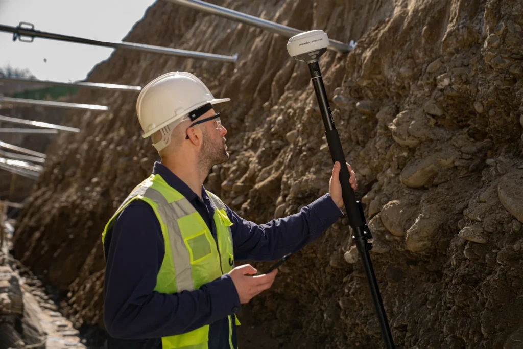 Construction surveyor in a safety vest and hard hat using a GNSS receiver to measure an exposed utility trench before backfill. The image fits the article’s focus on capturing accurate as-built field data before the trench is closed.