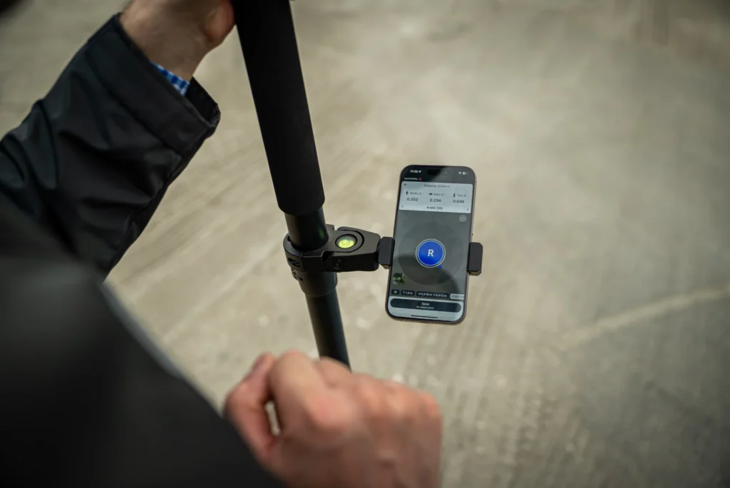 A person holds a surveying pole with a mounted smartphone displaying a surveying or mapping app, while working outdoors on a concrete surface.