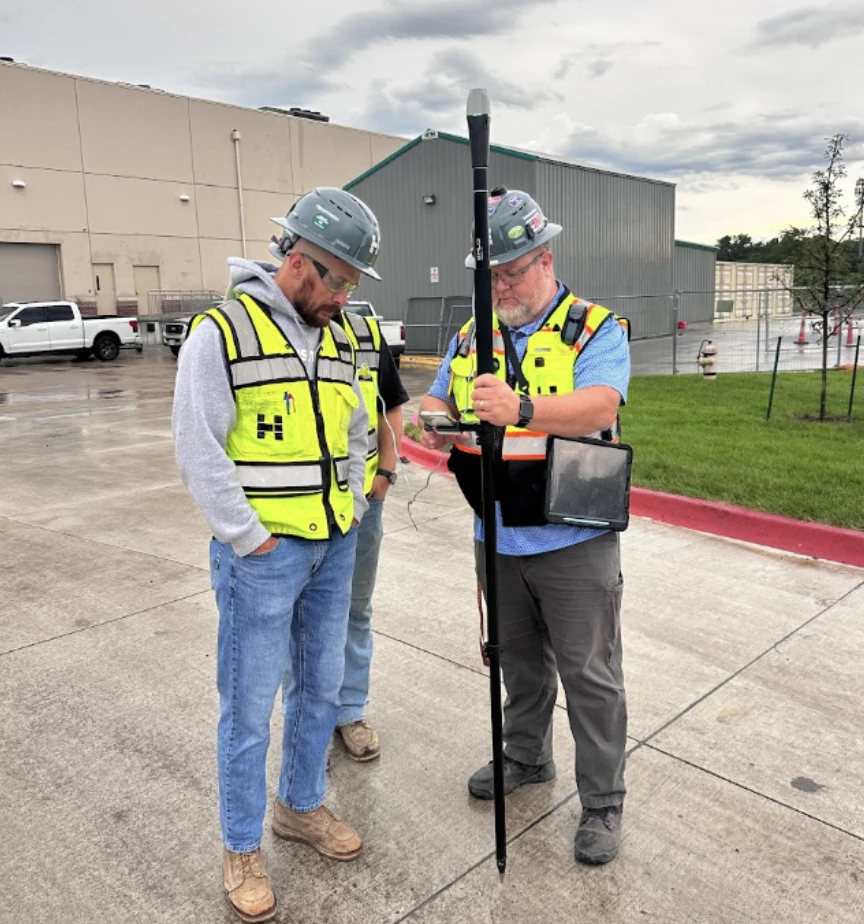 Two construction professionals wearing hard hats and high-visibility safety vests stand outdoors beside an industrial building. One man holds a surveying pole and measurement equipment while the other looks on. The scene includes a concrete driveway, parked trucks, a grassy area, and overcast skies.