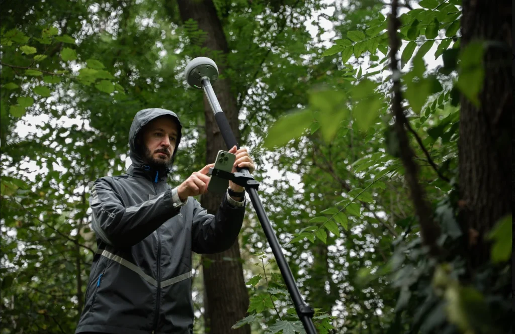 A surveyor in a rain jacket holds a Reach RX2 GNSS rover on a pole overhead while standing on a wooded trail under dense tree canopy.